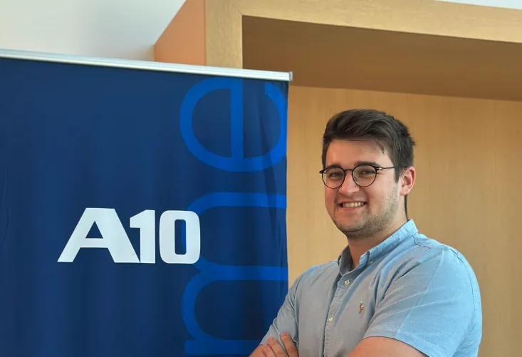 Ozgur Tufekci next to a blue A10 banner, wearing glasses and a light blue shirt, arms crossed.