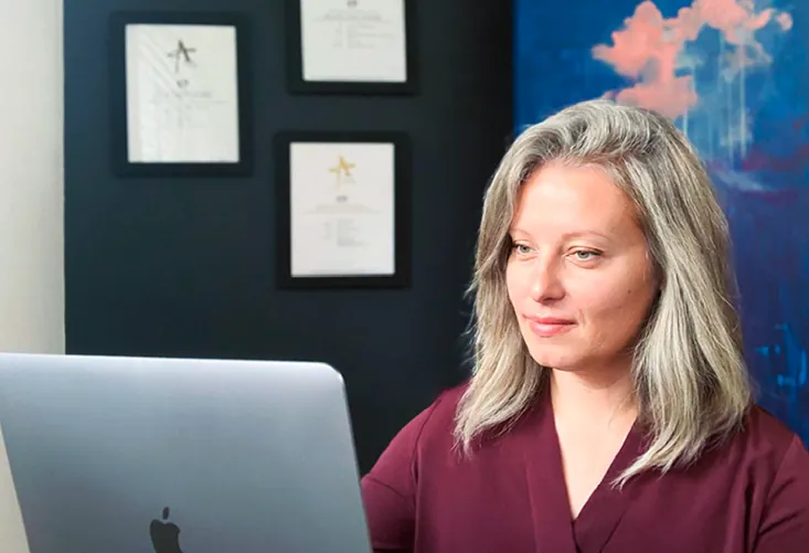 Woman working on a MacBook in a modern office with framed certificates on the wall.