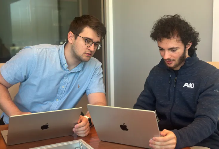 Picture of two teammates sitting at a table looking at a computer.