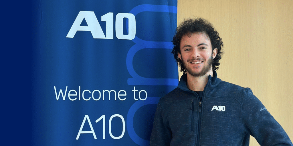 Man smiling next to a banner reading Welcome to A10 wearing an A10 jacket.
