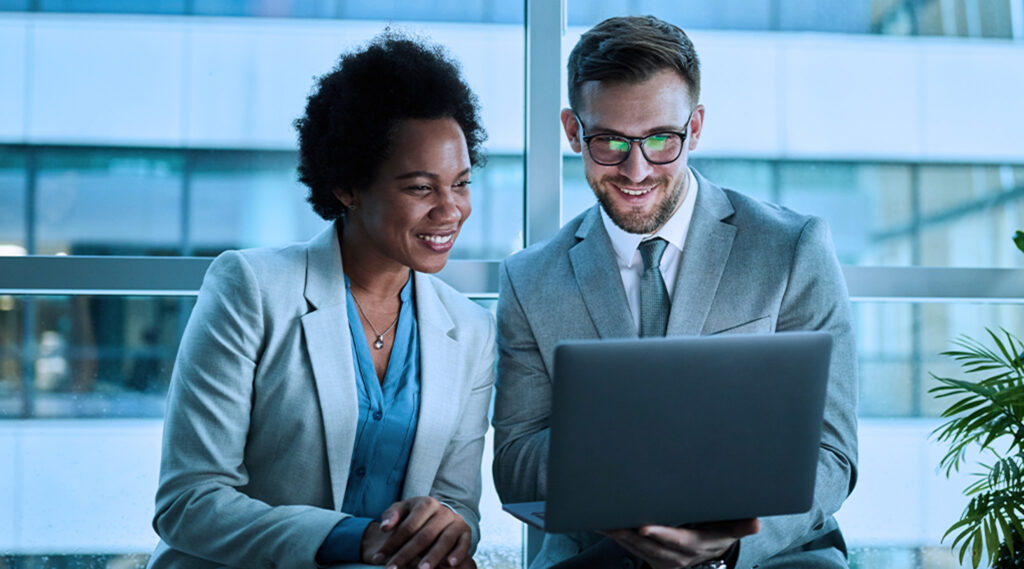 Two business professionals smiling while looking at a laptop in a modern office setting.