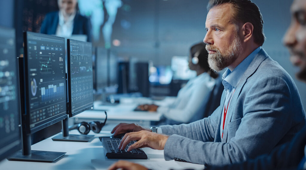 Man in a blue suit working on multiple computer screens displaying graphs and data in a modern office setting.